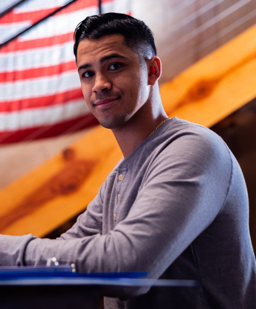 person in long sleeve shirt sitting at desk with American flag behind