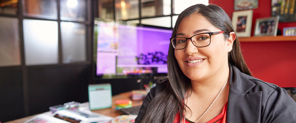 A woman with glasses sits at her desk, her computer in the background