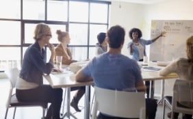 conference table with five individuals sitting around as woman at front is pointing at whiteboard and talking with those at table