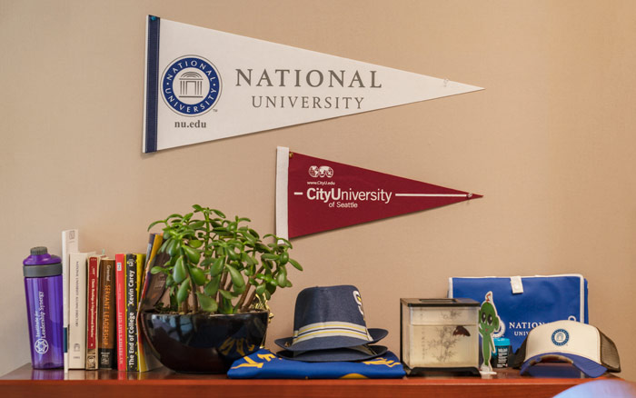 Nationa University and City University pennants are pinned about a shelf of books, plants, and alumni merchandise