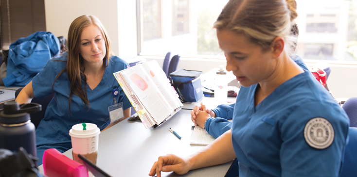 two women in scrubs are in a meeting, sitting across each other at a desk