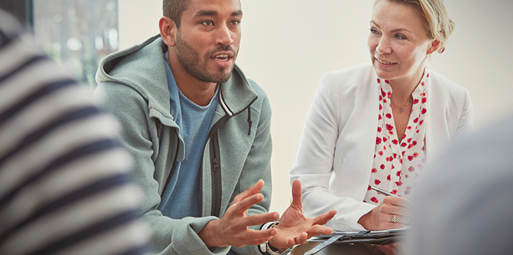 Man in a hoodie sweatshirt talks to a group of people sitting at a table. A woman listens to his left.