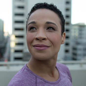 a woman with short curly hair looks up and out of frame in front of high rise buildings