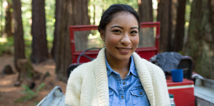 woman in a sweater stands in front of a truck parked in front of redwood trees