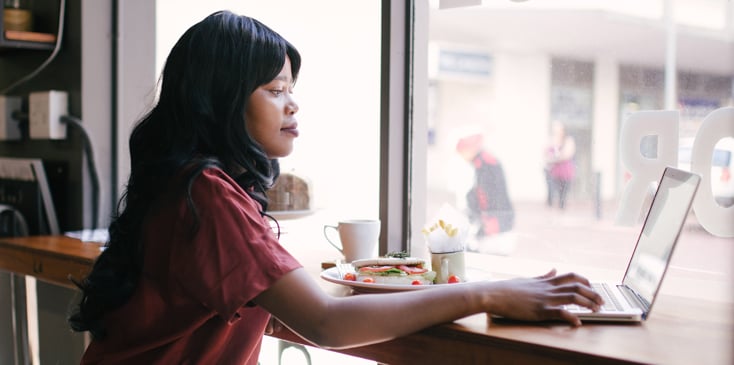 A woman sits at a cafe window with a sandwich and her laptop