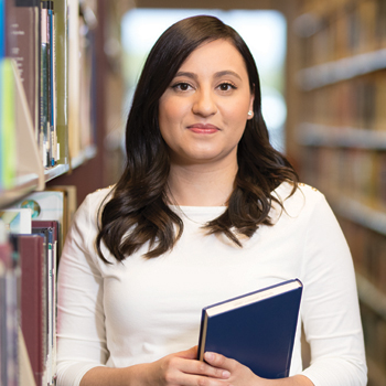 A woman in a white shirt stands with a blue book in between library stacks