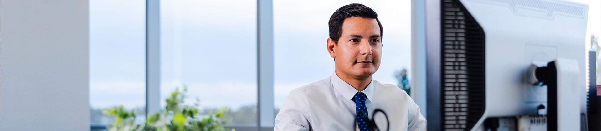 Andres C., a man in a white dress shirt and blue tie, stands in front of a window and looks off to the side.