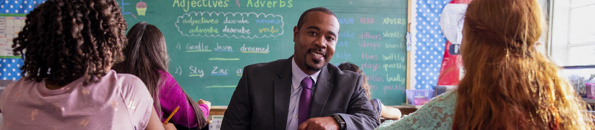 Prince M. sits in a classroom of school children. He is wearing a dark suit and purple tie.