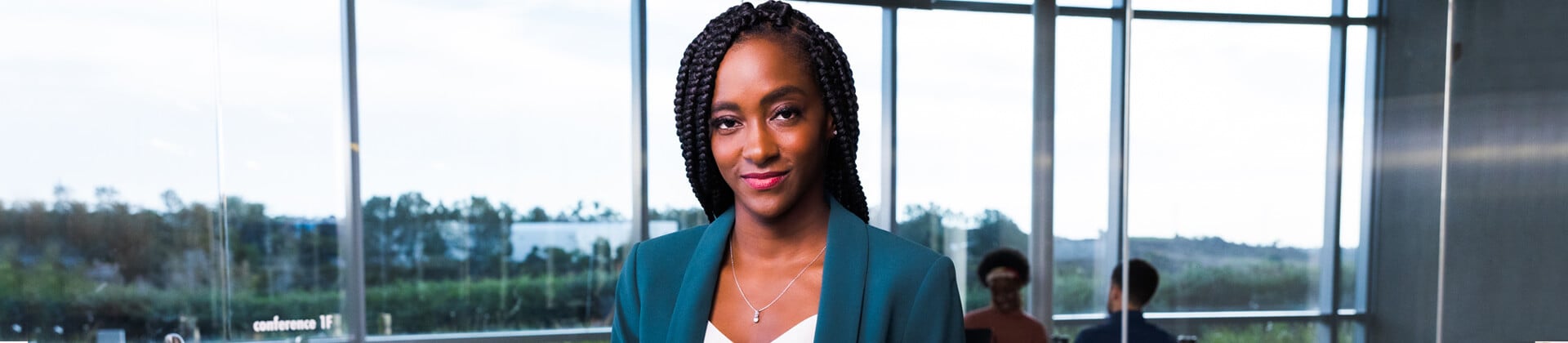 Spshelle R. stands in front of a large glass window. She wears a teal blazer and long box braids