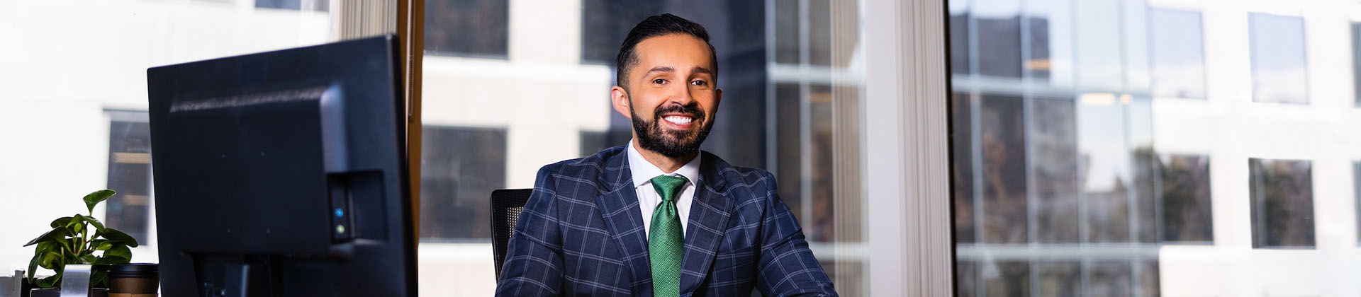 Ivan F. sits at a desk with a computer. He is wearing a navy blue plaid suit with a green tie