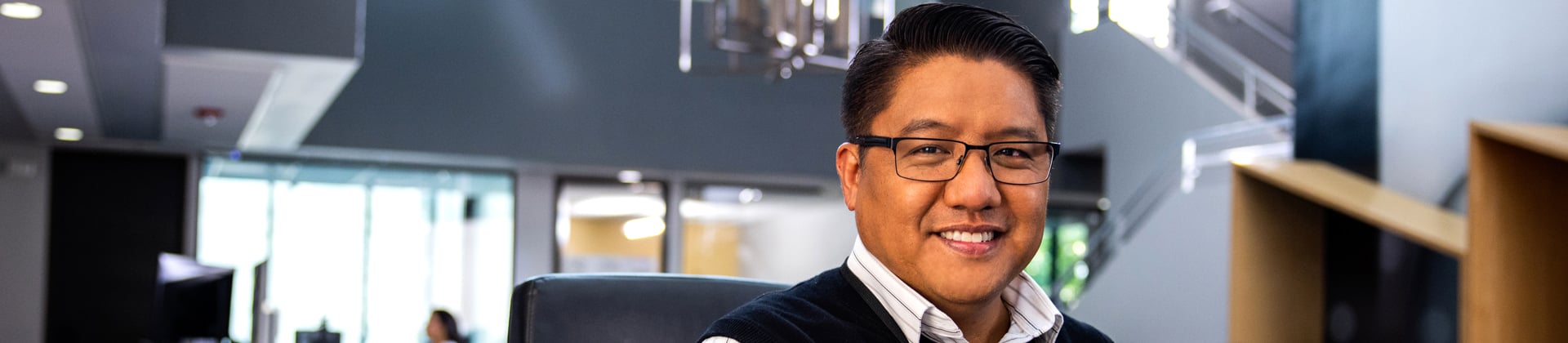 Edward H. sits in a chair in a modern office. He wears glasses and a navy blue sweatervest.