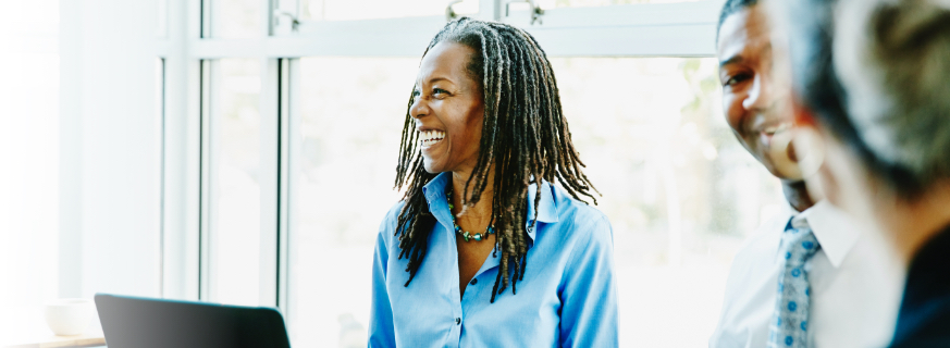 Professional woman with microlocs stands in front of a laptop as colleagues talk near her