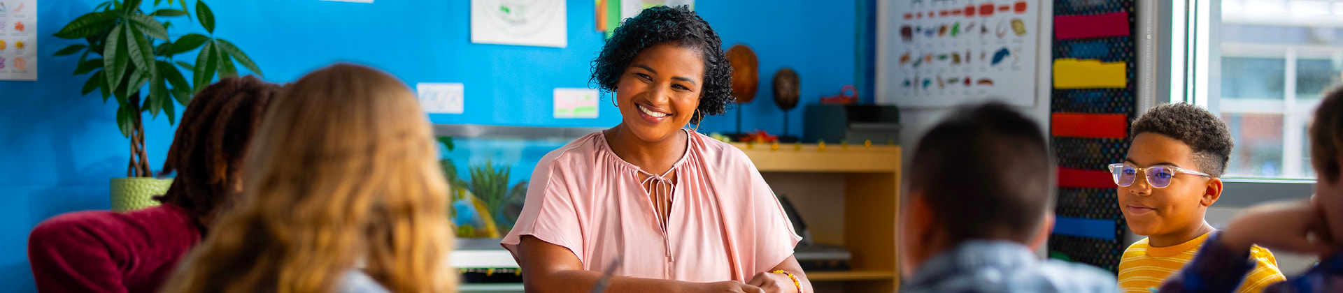 Toya S., a woman with short curly hair and a pink blouse, teaches a group of students in a classroom