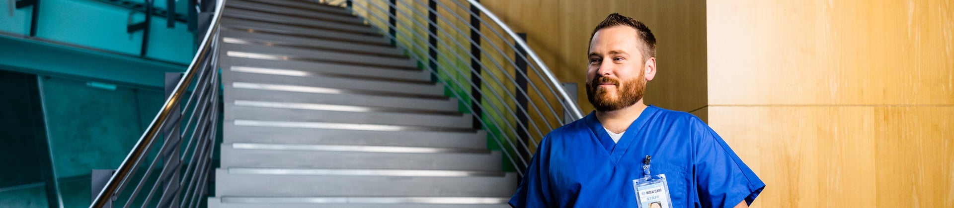 Kyle W., Class of 2018, looks past the camera. He is standing in front of a staircase and is wearing blue scrubs.