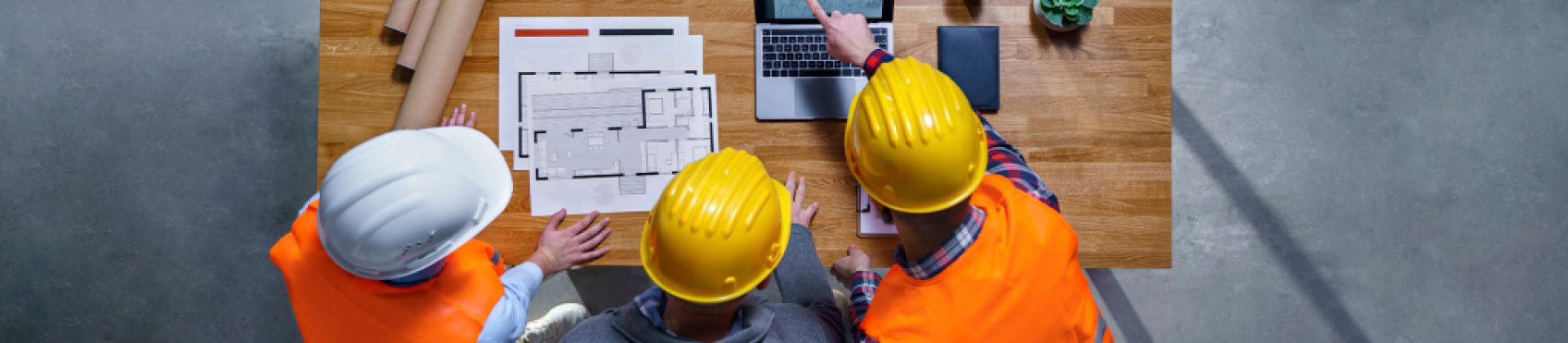 three construction workers in hardhats and safety vests look over blueprints of a building.