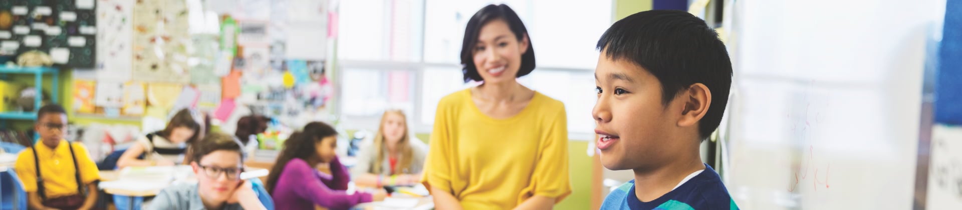 A woman in a yellow blouse looks at a young student who is in the foreground of a classroom