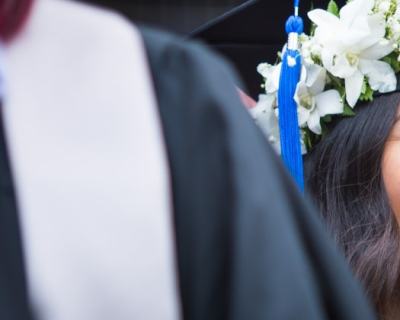 woman in cap and gown smiling off to her left wearing flowers around graduation cap after earning her bachelor's degree