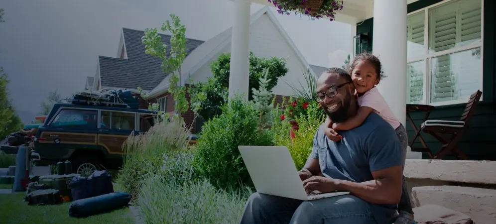 Kristian E., Class of 2018 black man sitting outside on laptop as toddler child wraps arms around his neck from behind