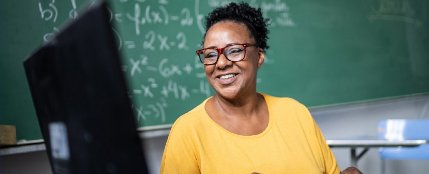 teacher in yellow shirt at desk and chalkboard behind them