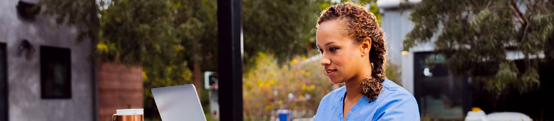 Woman in blue scrubs with braided curly hair sits outside looking at a laptop