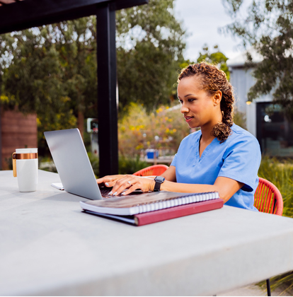 A woman in scrubs works outside on a laptop