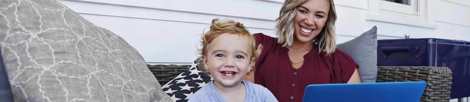 A mother sits on a couch with a laptop and smiles at her toddler-aged son, who is looking forward.