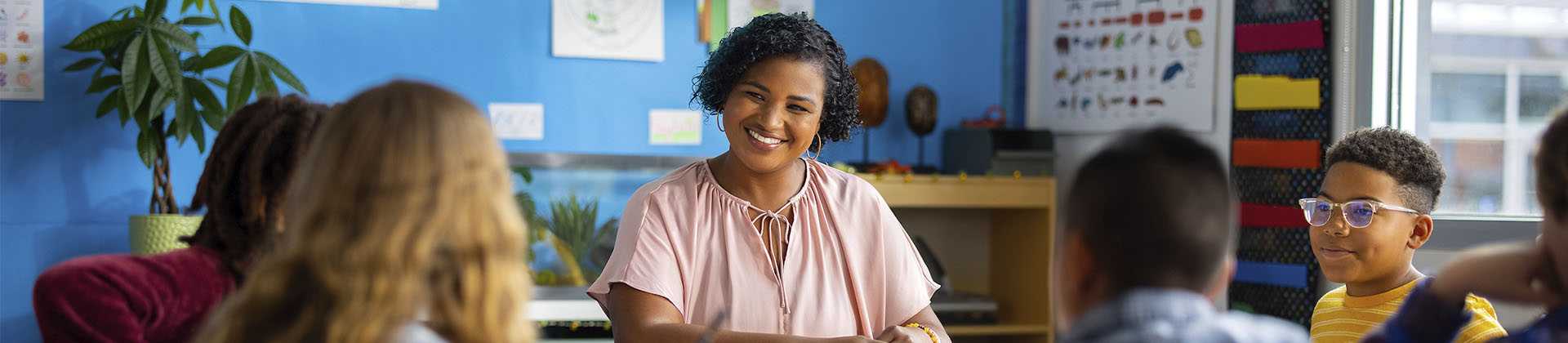 Woman in light pink blouse smiles at a group of elementary school students in a classroom.