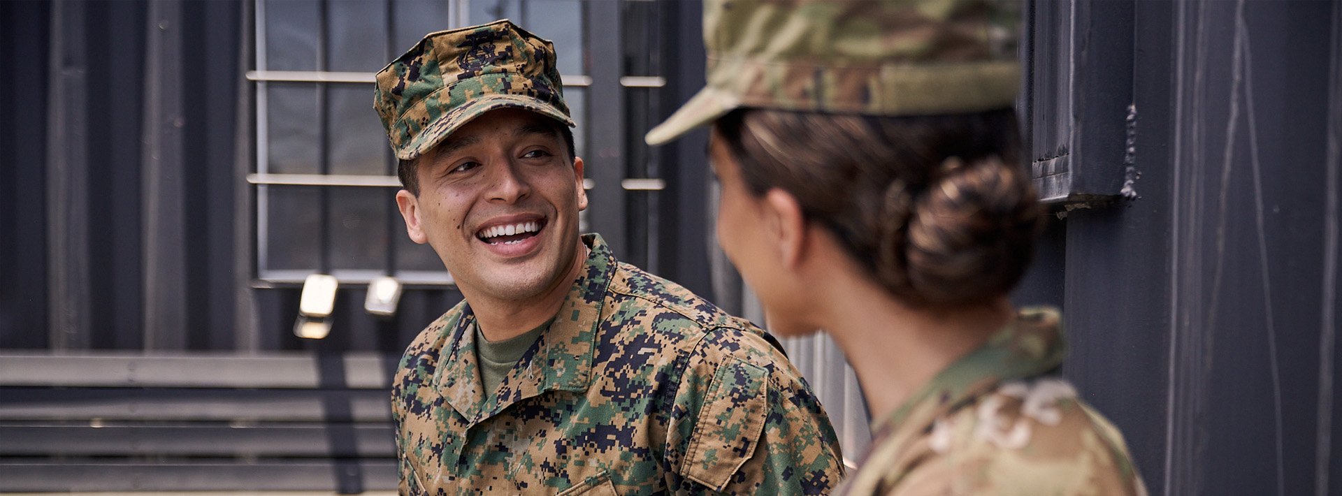 two military servicemembers in uniform talk next to a gray building