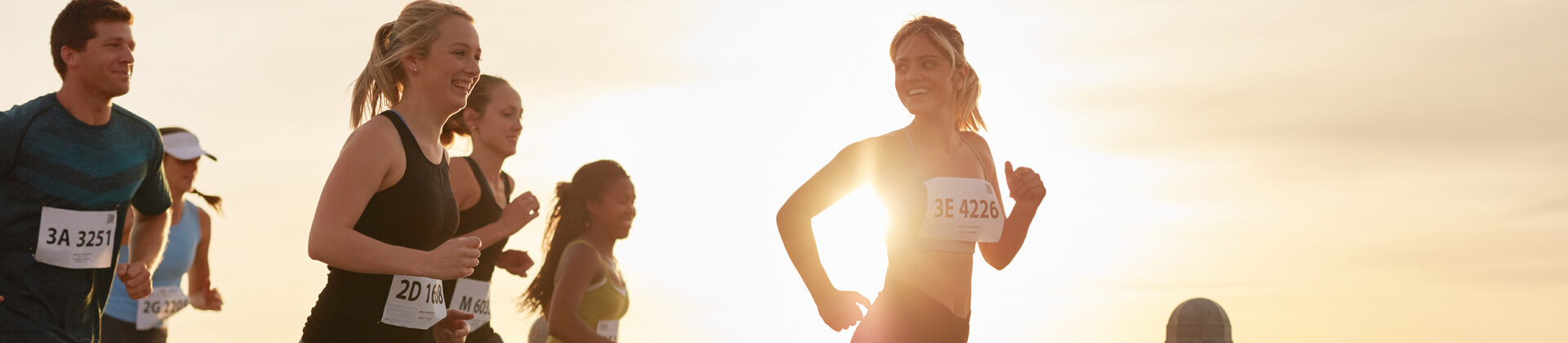 Women in tank tops and numbers run a race and smile with the sun behind them 
