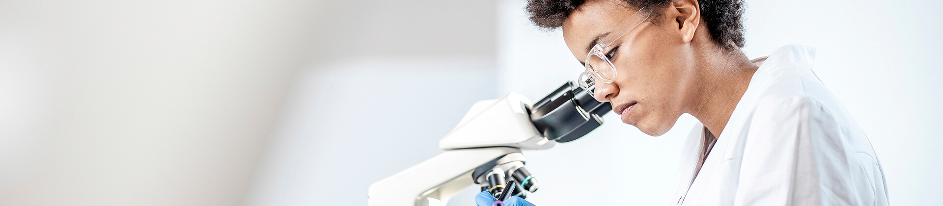 A woman with short, coiled hair and glasses looks down into a microscope for the Bachelor of Science in Clinical Laboratory Science Program Page