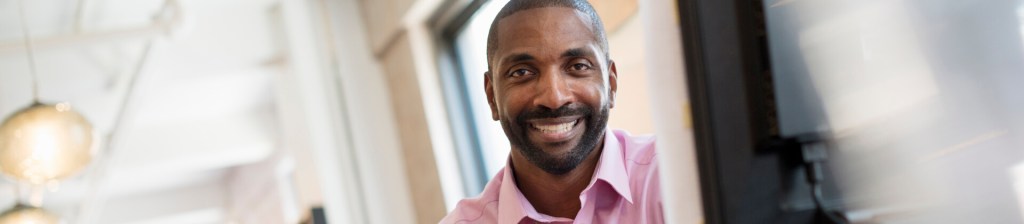 man sitting by computer monitor smiling into camera