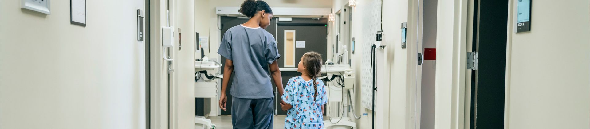 A woman in scrubs looks down at a young girl patient while they walk down a hospital hall