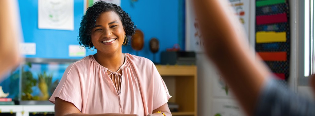 teacher in classroom smiling as students have hands raised