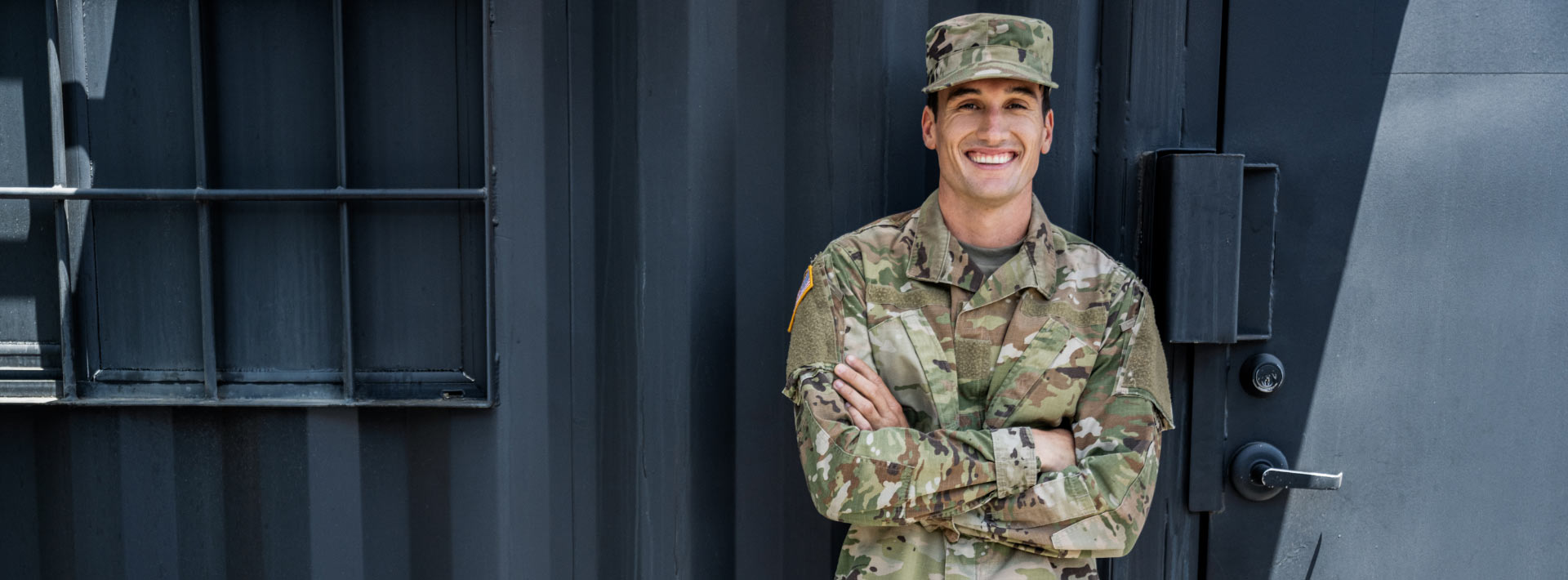 Man in an army uniform stands with his arms crossed in front of a building