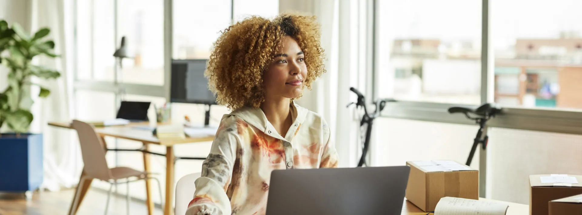 A woman with blonde curly hair works on a laptop and looks off into the distance