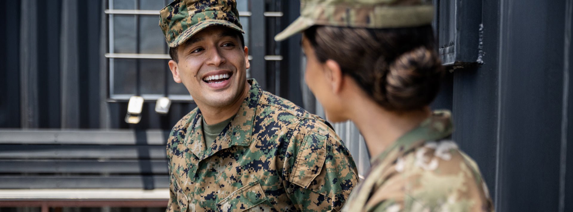 A man and a woman, both in camo uniforms, talk in front of a building