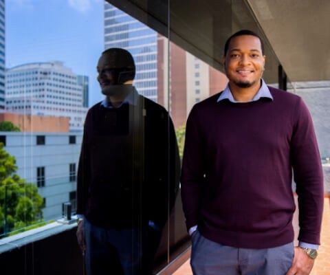 A man in a purple sweater stands next to a reflective window