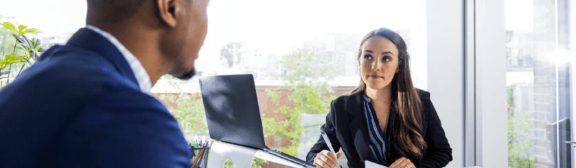 woman in office looking at man while she holds pen in hand and paper in the other