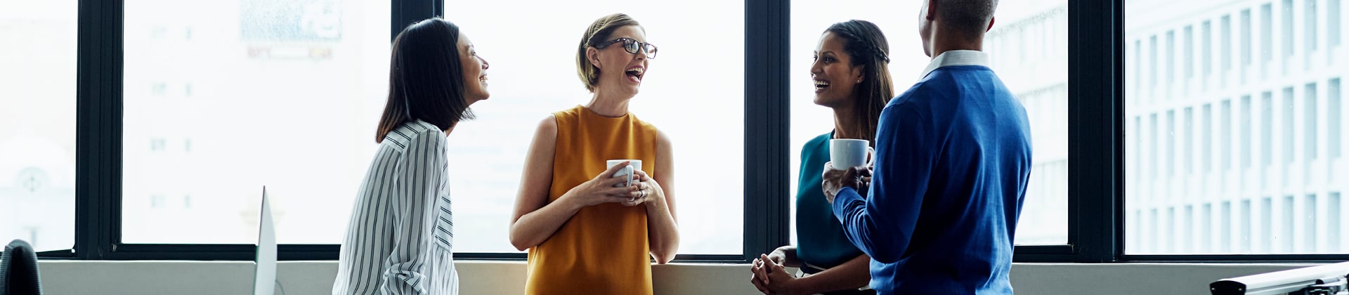 3 woman and one man standing with coffee mugs engaged in conversation and smiling 