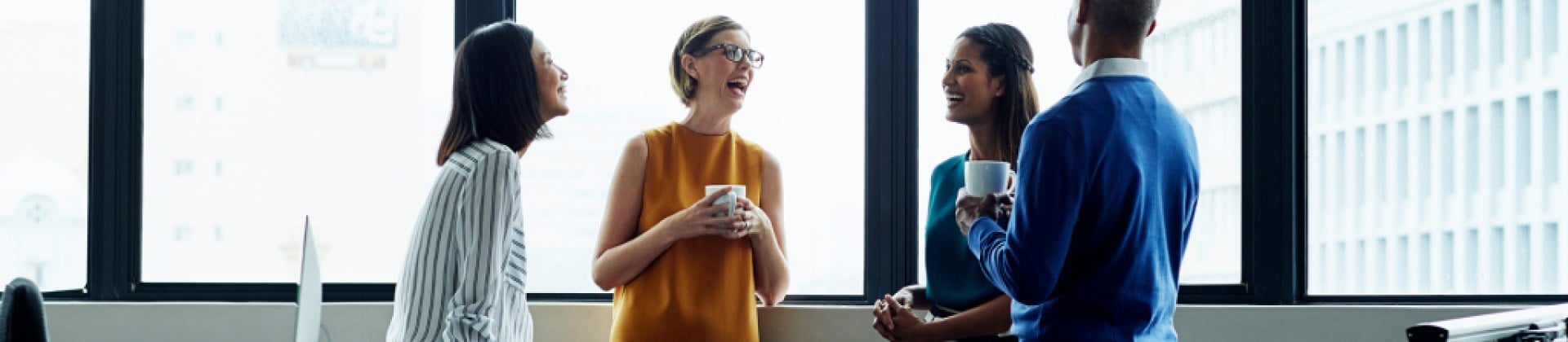 four individuals in a semicircle engaged in conversation, two of them with coffee mugs, three women are smiling, one man has back turned to camera