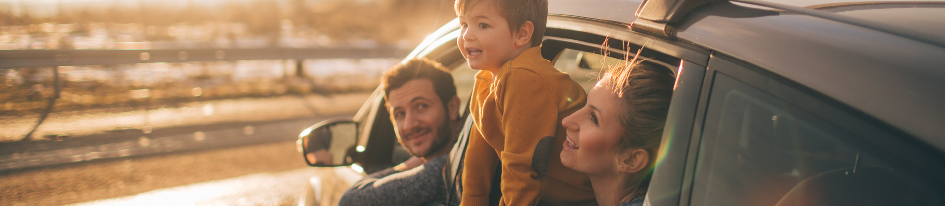 a family with the man in drivers seat with woman and preschool aged boy on her lap - all leaning out of the window - the father looks at the boy smiling, the mother and boy are looking at something in the distance, smiling. The sun is glowing on them. 