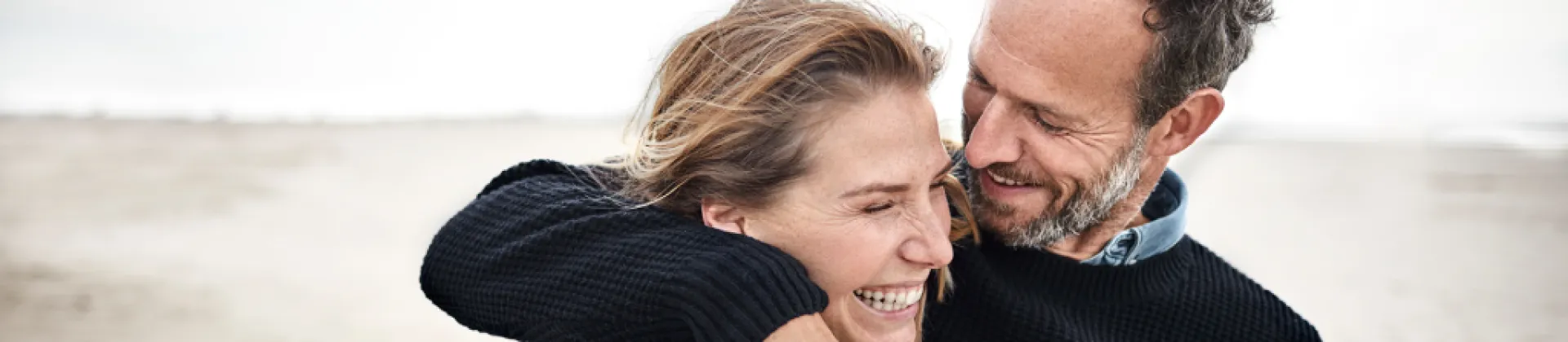 later middle aged couple smiling at the beach, woman's eyes are closed and man is looking down at her embracing her