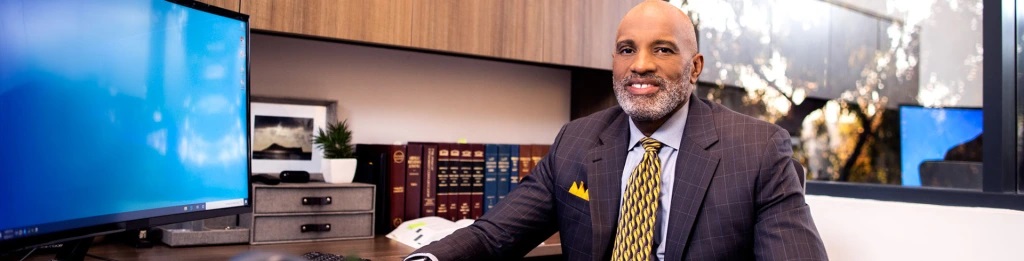 man in suit at desk in office with books behind him and desktop to his right, smiling into camera