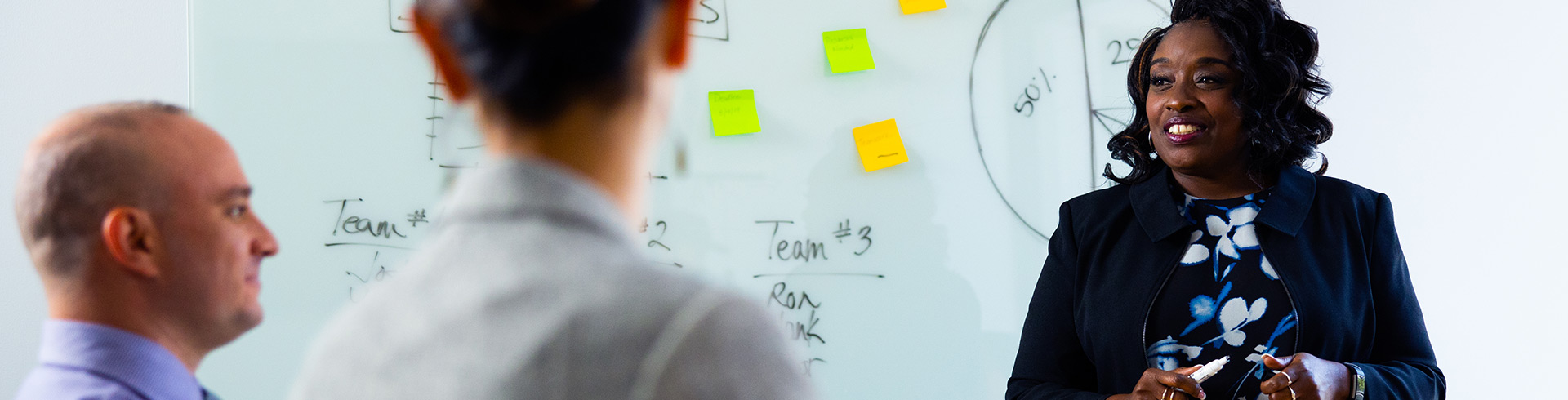 woman in a blazer teaches a few men in front of a white board