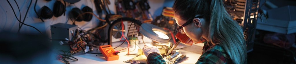 woman with glasses and ponytail soldering a circuit board