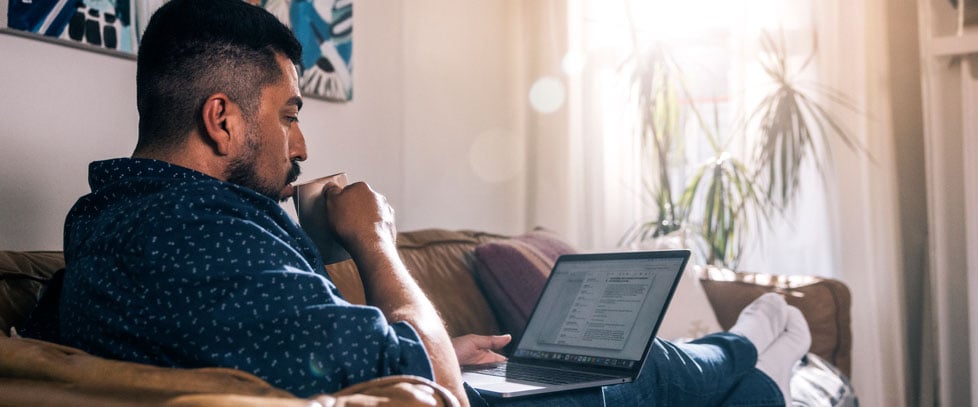 man sips tea while working on a laptop on his couch