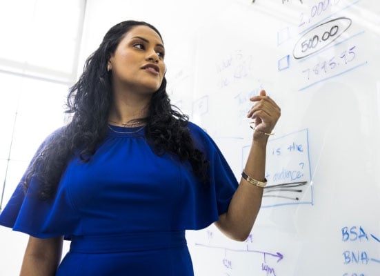 a woman in a blue top stands teaching at a whiteboard