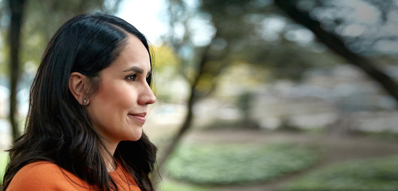 Abigail wears an orange shirt and stands outside in front of some trees. She has ear piercings and long dark hair.