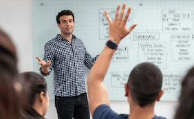 person teaching in front of whiteboard and student raising their hand
