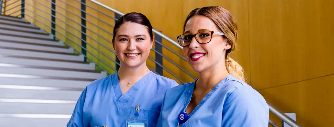 two nurses in blue scrubs standing at bottom of stairs
